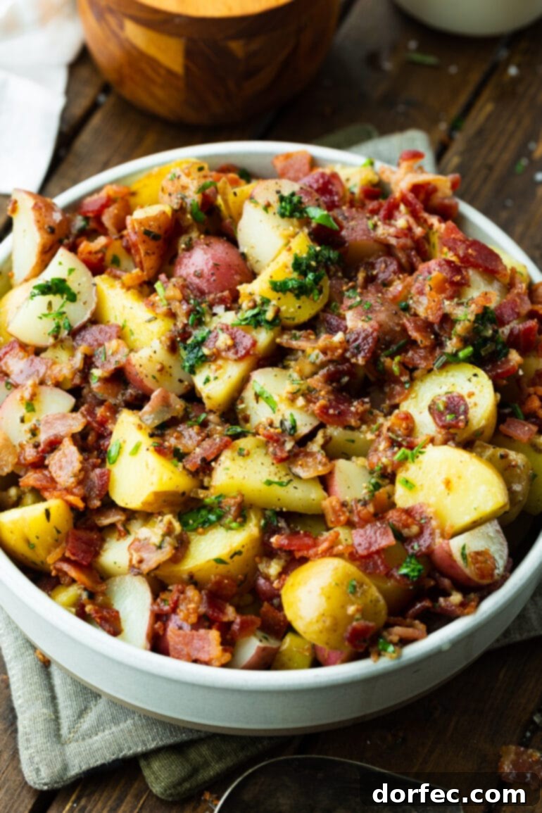 a photo of a large white bowl full of German potato salad consisting of cubed potatoes, bacon bits and parsley.