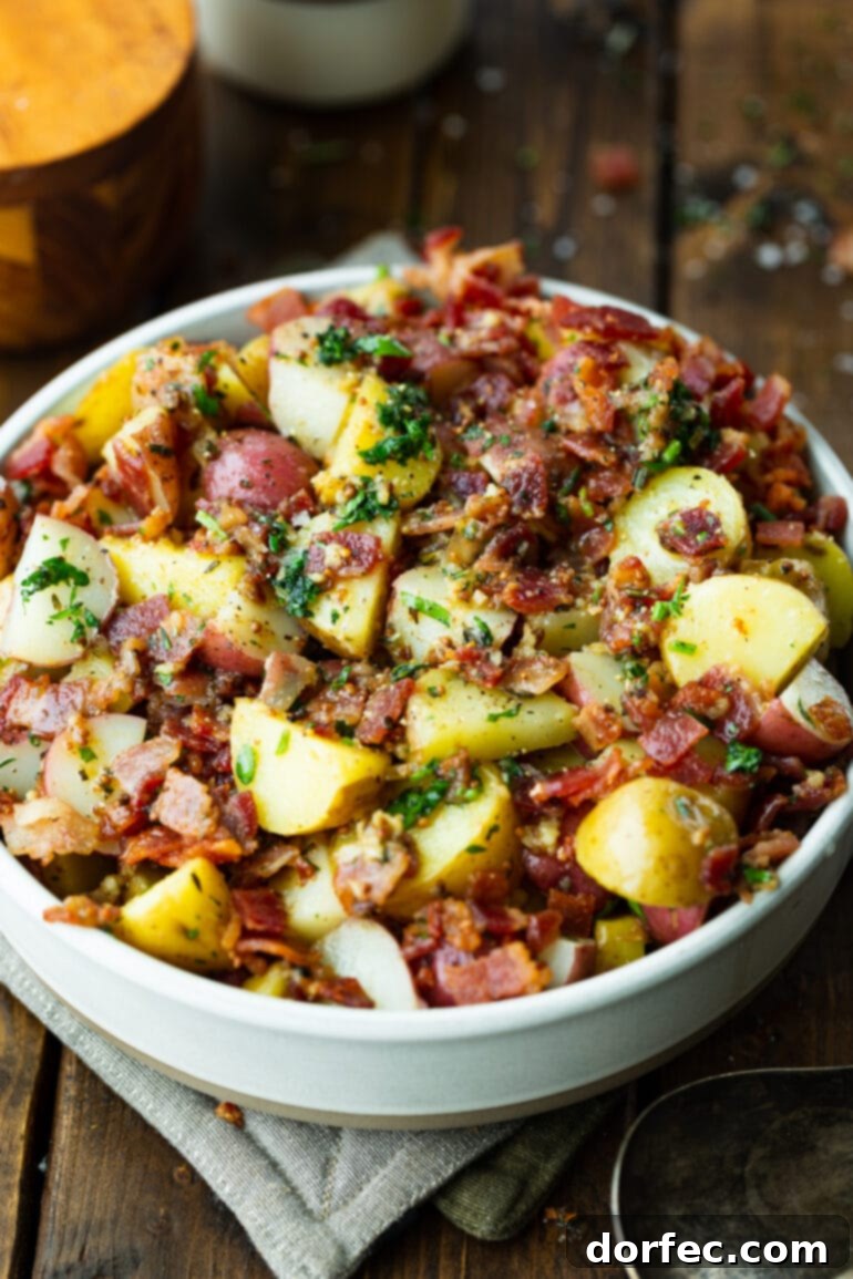 a photo of German potato salad consisting of cooked potatoes, bacon crumbles, garlic and parsley in a large white serving bowl.