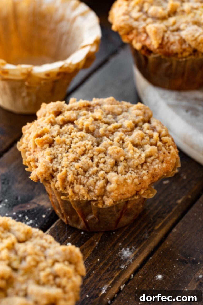 Ultimate Streusel Banana Bread Muffins 4 A single, perfectly golden-brown banana crumb muffin sits on a rustic wooden table, its paper liner partially peeled away, with other blurred muffins in the background.