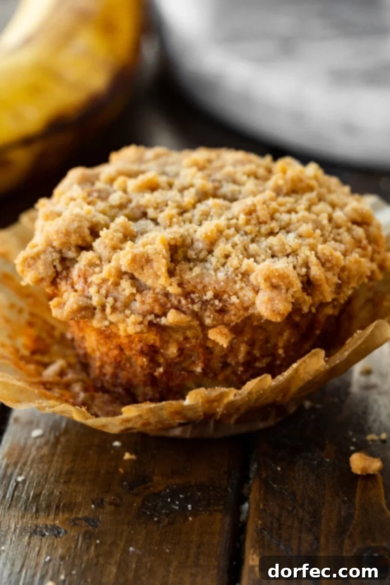 Ultimate Streusel Banana Bread Muffins 6 A single banana muffin with a generous crumb topping, presented in its muffin liner which is gently pulled away from the sides. The focus is on the inviting texture and golden-brown bake.