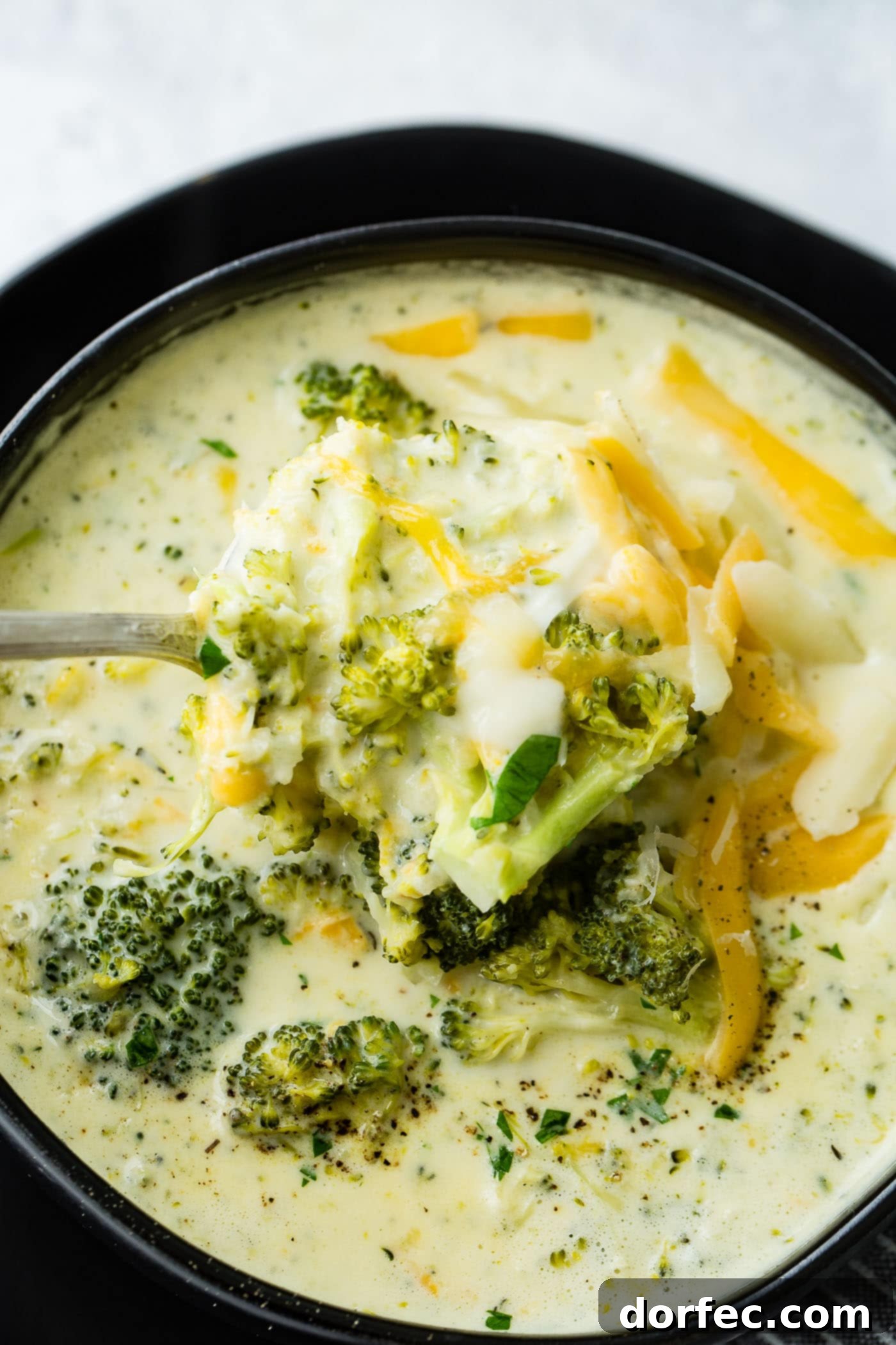 A steaming black bowl filled with creamy slow cooker broccoli cheese soup, generously topped with extra melted cheese and fresh green herbs.