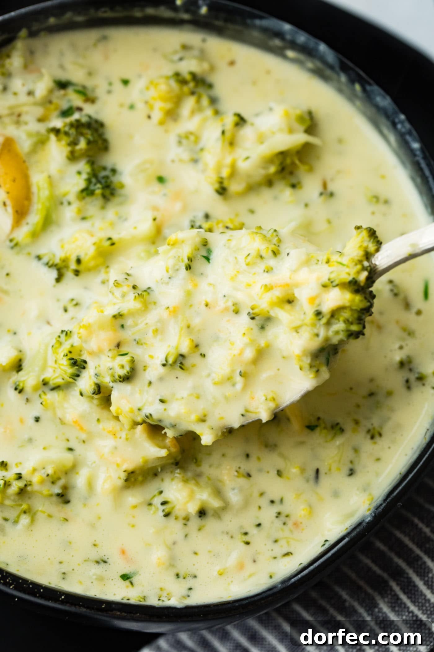 A close-up shot of a black bowl brimming with luscious slow cooker broccoli cheese soup, adorned with a generous topping of shredded cheese and a sprig of fresh thyme.