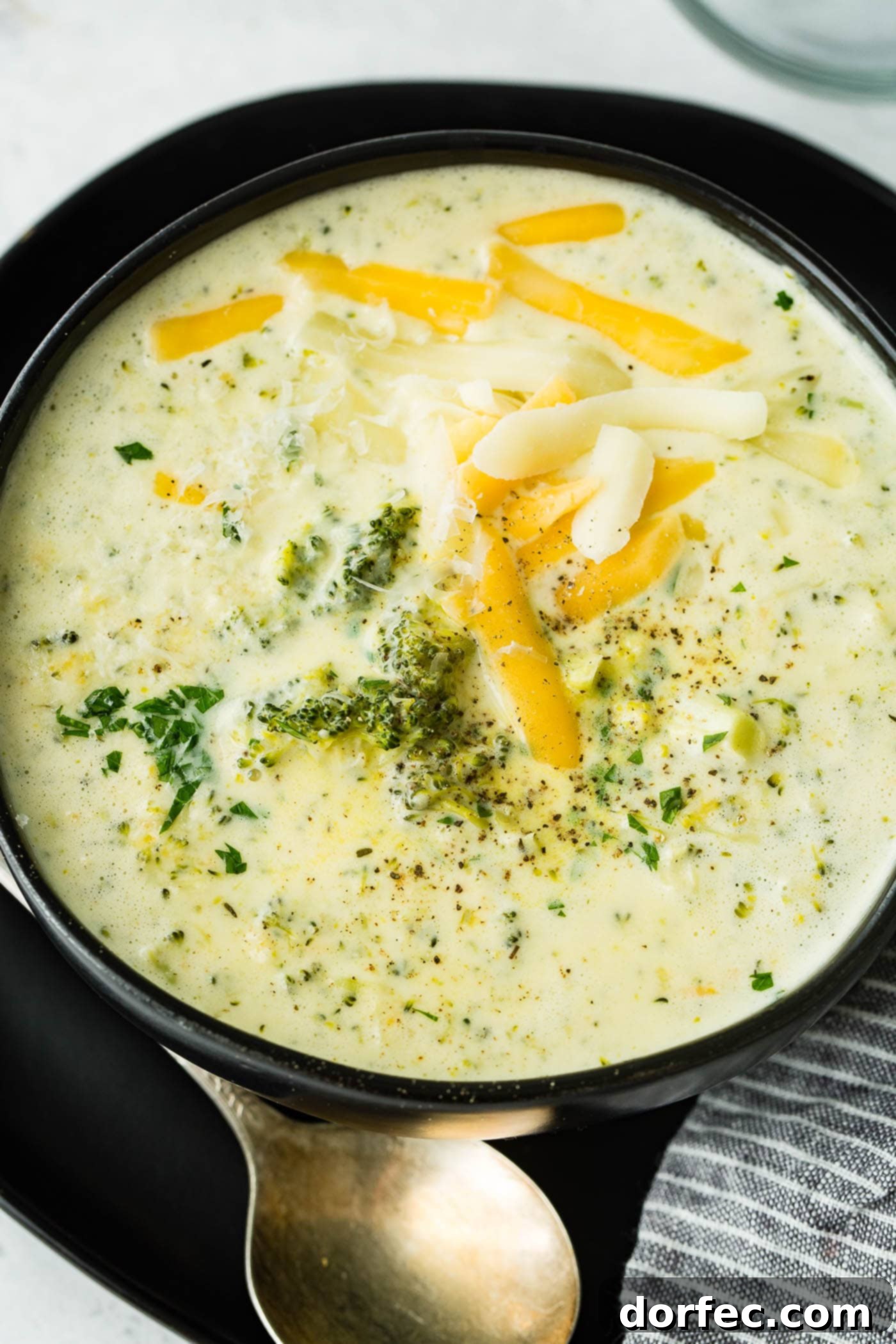 A black bowl of slow cooker broccoli cheese soup, exquisitely presented with a generous topping of shredded cheese and a sprig of fresh rosemary for garnish.