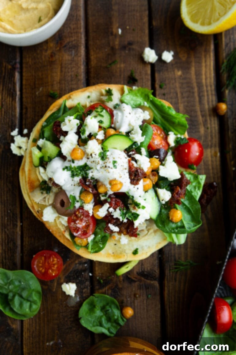 A photo taken over the top of a Greek chicken tostada topped with cherry tomatoes, lettuce, olives, roasted chickpeas, cucumbers, and tzatziki sauce, showcasing a colorful and healthy meal.
