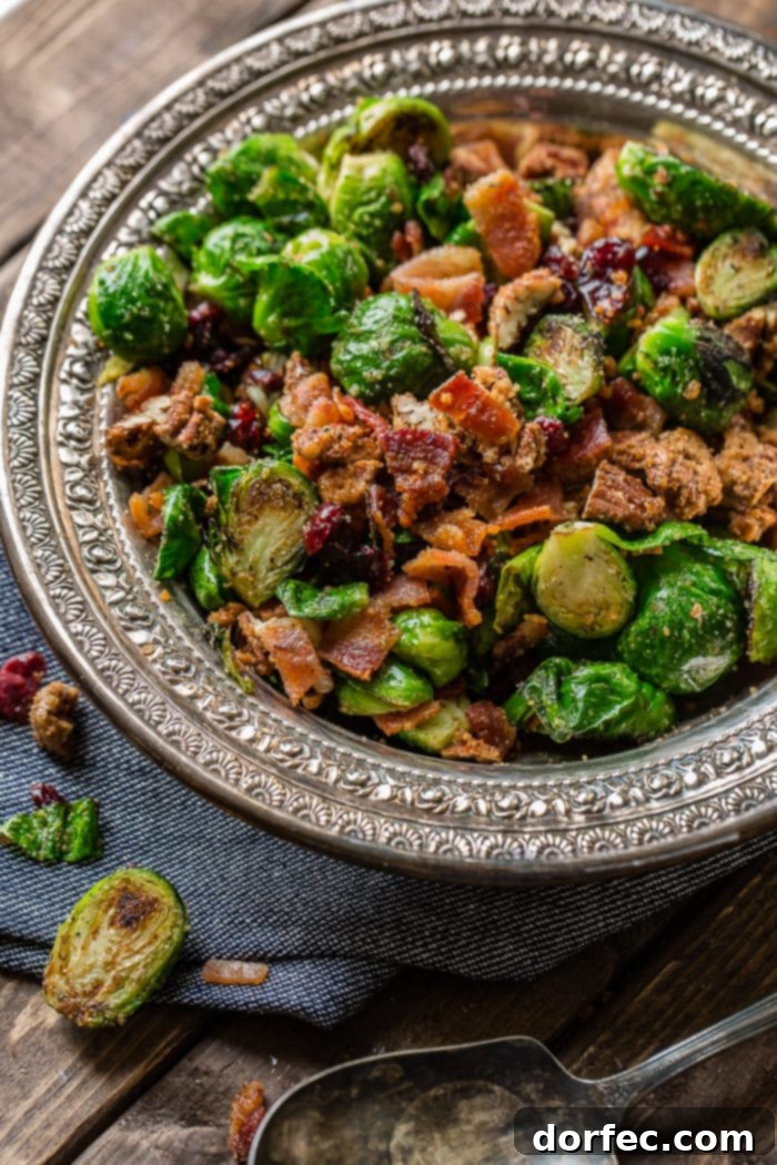 A close-up shot of a vintage silver bowl filled with vibrant green Brussels sprouts, halved and pan-roasted until tender with golden-brown edges. They are generously mixed with crimson craisins, savory crisp bacon, and glistening candied pecans, resting on a dark denim napkin.