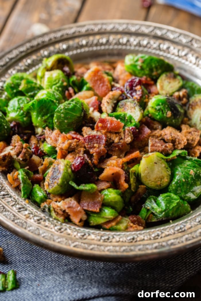 A beautiful close-up of the Pan Roasted Candied Pecan Bacon Brussels Sprouts, showcasing the crispy bacon, glazed pecans, and tender, golden-brown sprouts in a silver serving bowl.