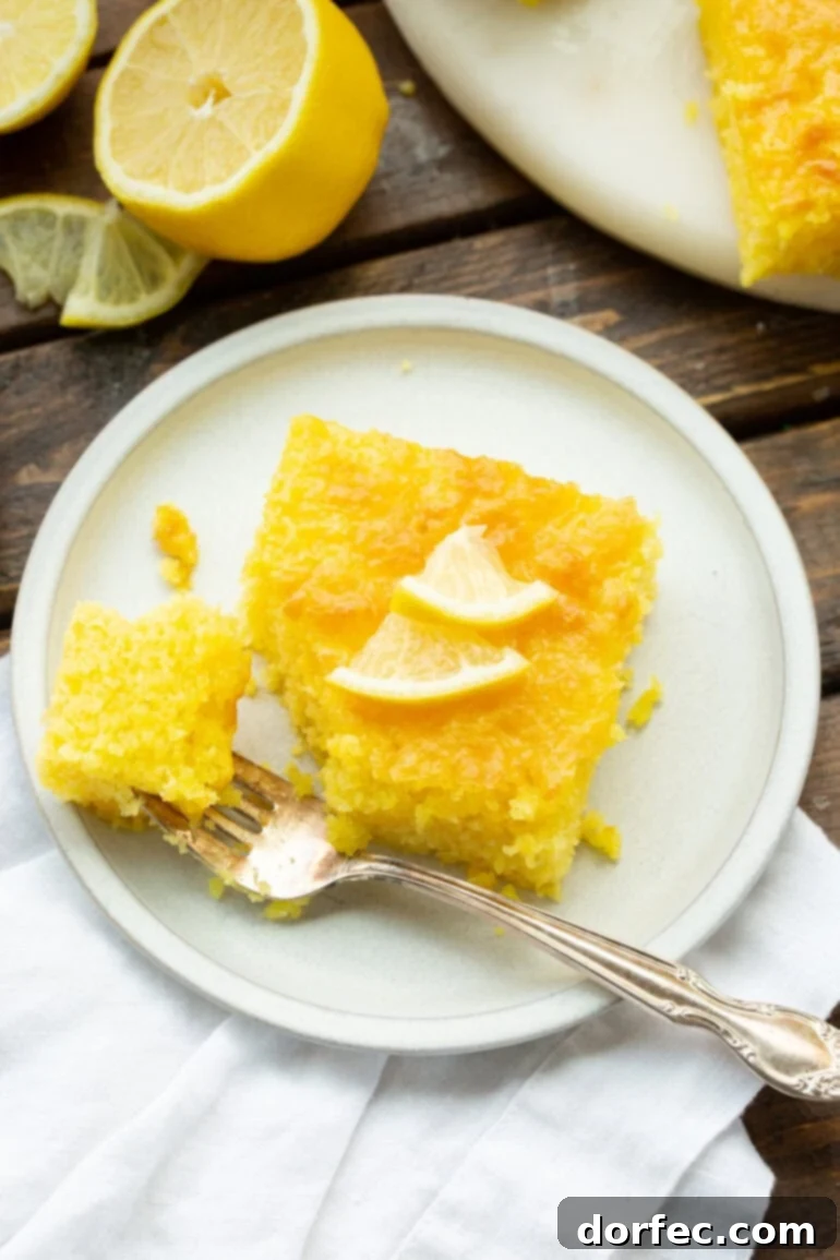 A slice of moist Lemon Jello Cake on a white dessert plate, with a fork removing a bite, showcasing its tender texture.