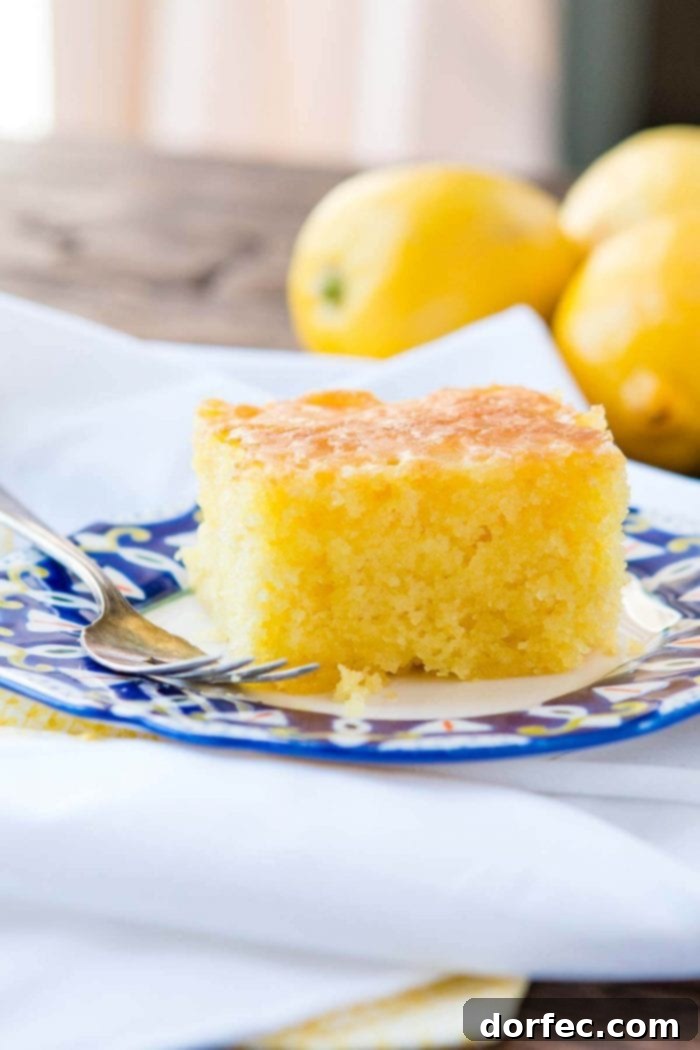 A delicious slice of Lemon Jello Cake on a decorative white plate, with a silver fork ready to enjoy.