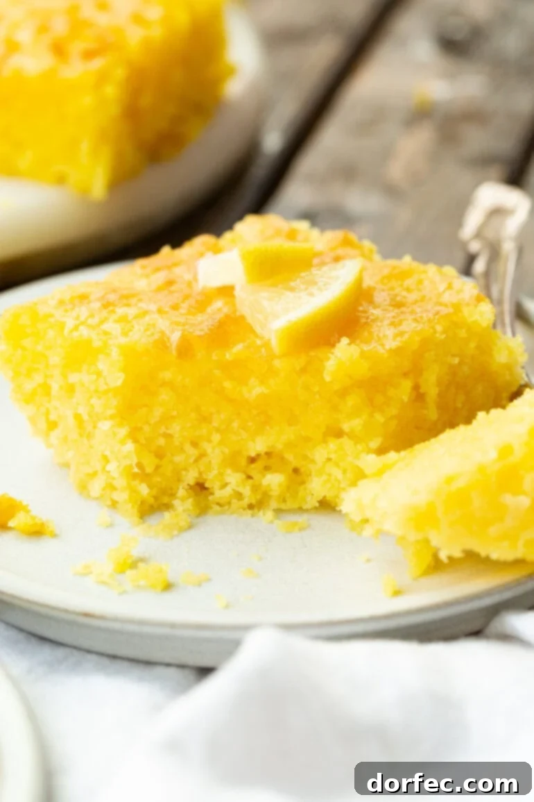 A close-up photo of a moist slice of Lemon Jello Cake with a bite removed, revealing the soft, bright yellow interior.