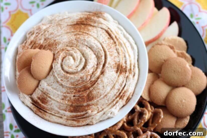 A vibrant Pumpkin Cream Cheese Dip served in a modern black bowl, artfully arranged on a dark plate with a colorful array of dippers including sliced red apples, golden vanilla wafers, and twisted pretzels, ready to be enjoyed.