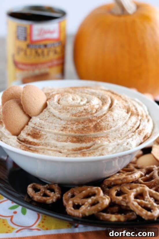 A close-up shot of a creamy Pumpkin Pie Dip in a white bowl, delicately sprinkled with extra pumpkin pie spice, surrounded by a rustic backdrop of pretzels and a decorative pumpkin, embodying the essence of fall.