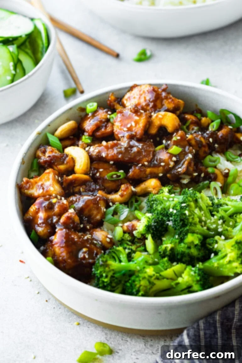 A close-up shot of a bowl filled with tender chicken pieces coated in a rich, dark sauce and mixed with roasted cashews, with steamed broccoli on the side.