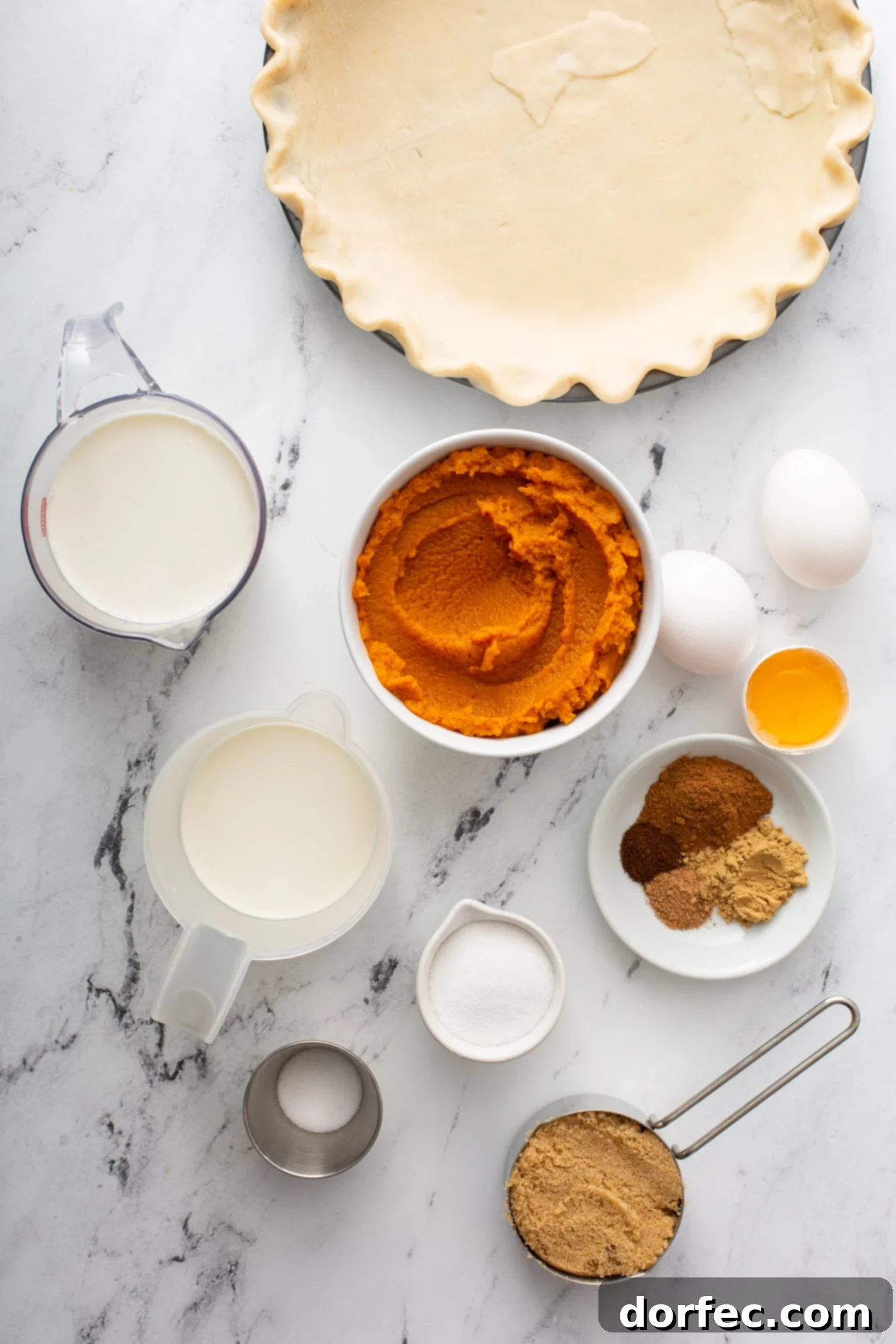 A collection of fresh, high-quality ingredients for a homemade pumpkin pie, including a can of pumpkin puree, heavy cream, eggs, and various fall spices, arranged artfully for baking.