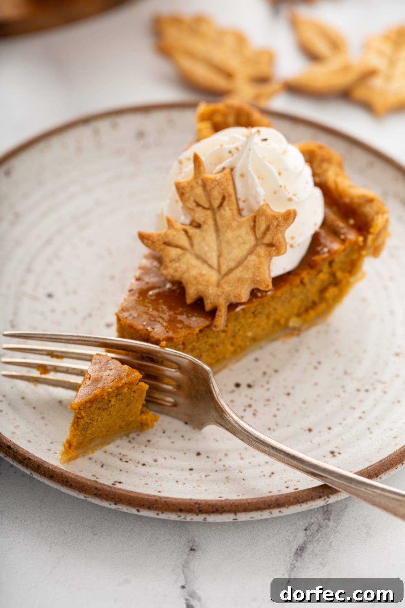 A fork is gently cutting into a creamy slice of golden pumpkin pie, beautifully topped with a swirl of piped whipped cream and a small, delicate pie crust leaf.