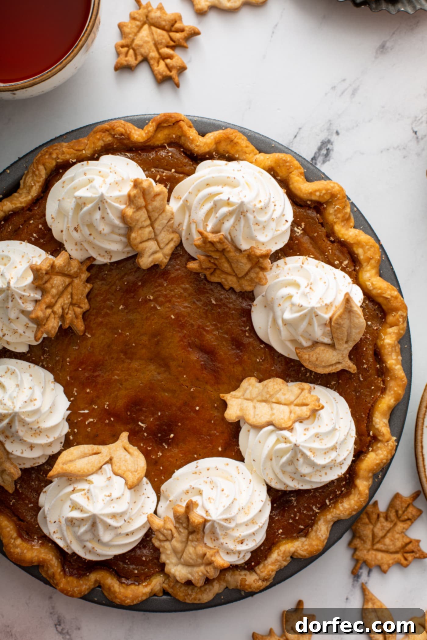 A stunning top-down view of a classic pumpkin pie, elegantly decorated with swirls of piped whipped cream and delicate, stamped pie crust leaves, showcasing an ideal fall dessert.