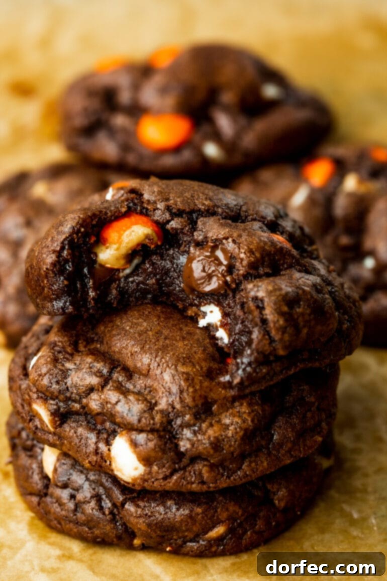A stack of three decadent Reese's Triple Chocolate Cookies, with a bite taken from the top cookie, showcasing their chewy texture and abundance of chips and Reese's Pieces.