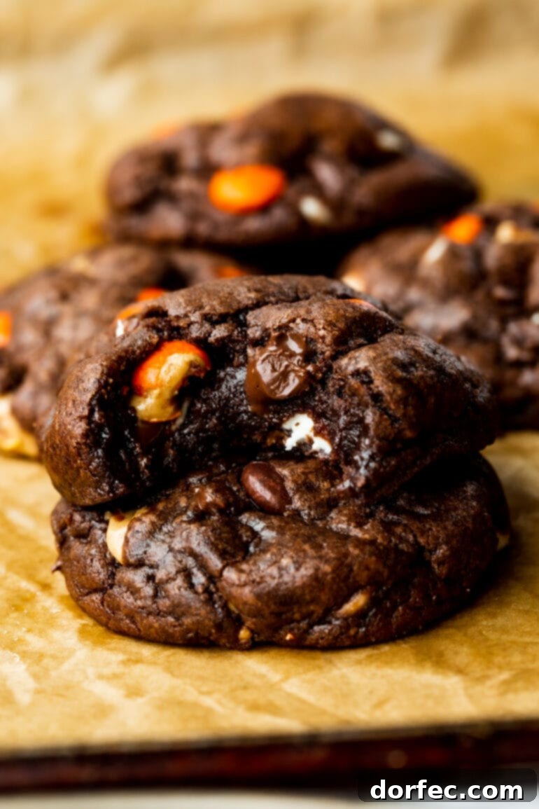 A close-up photo of thick and chewy triple chocolate cookies, one stacked on top of another with a bite taken out of the top cookie, revealing its rich, fudgy interior filled with chocolate and peanut butter pieces.