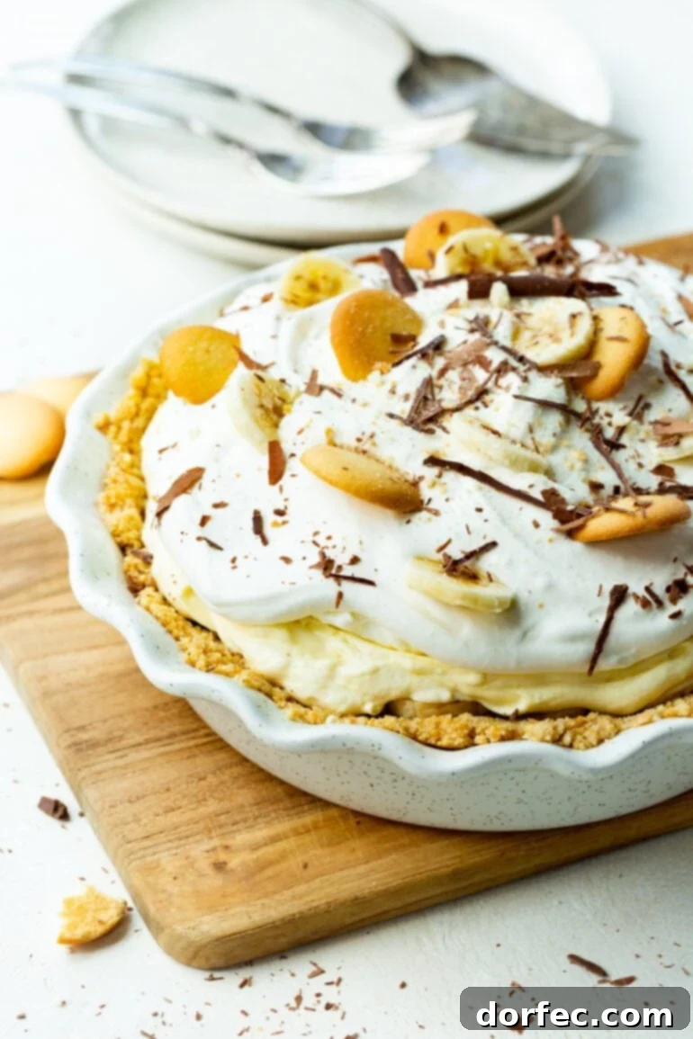 A photo of a whole banana pudding pie in a white pie dish topped with whipped cream, banana slices, Nilla wafers, and shaved chocolate