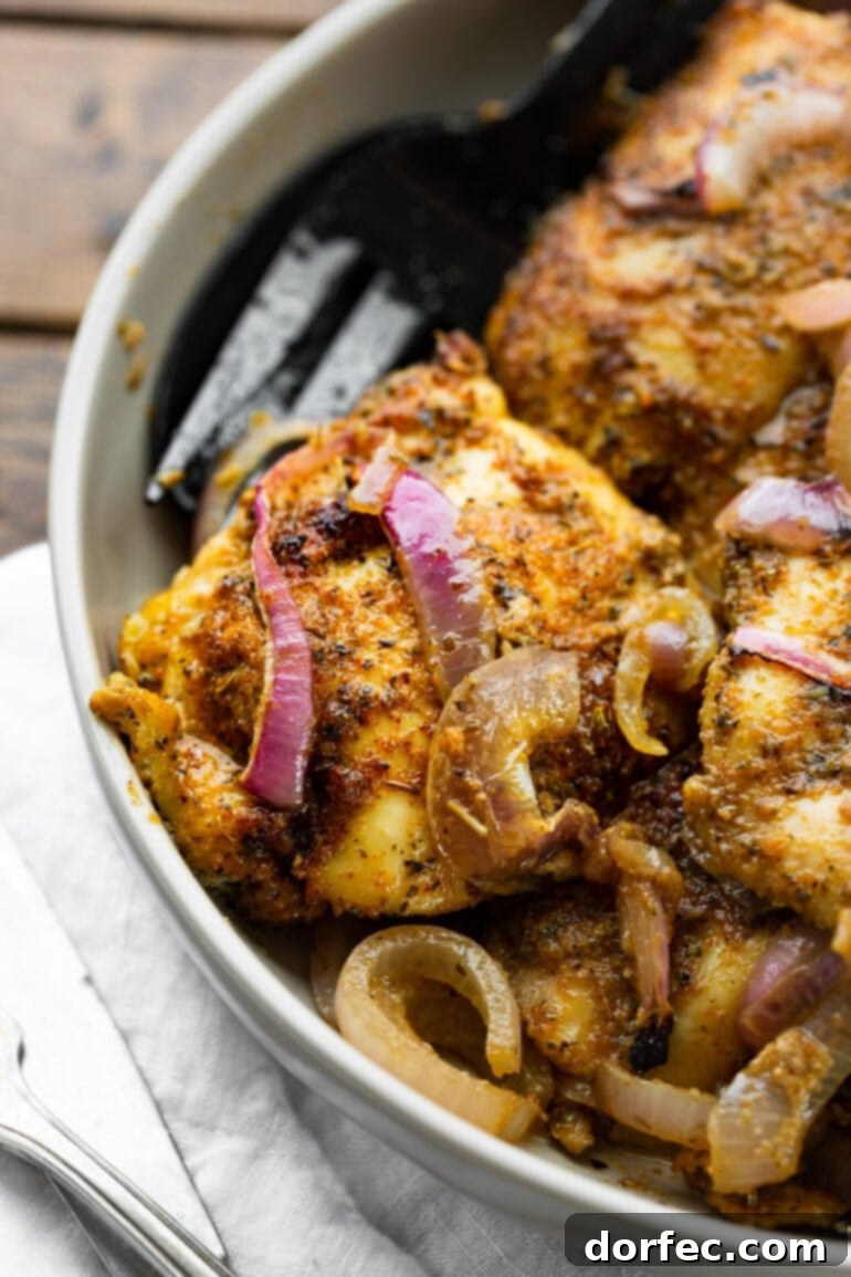 A close-up photo of a baked chicken thigh topped with slices of red onion and a delicious dry rub.