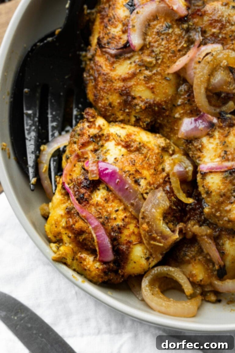 A close-up photo of a golden baked chicken thigh with a delicious dry rub, topped with cooked red onion pieces.
