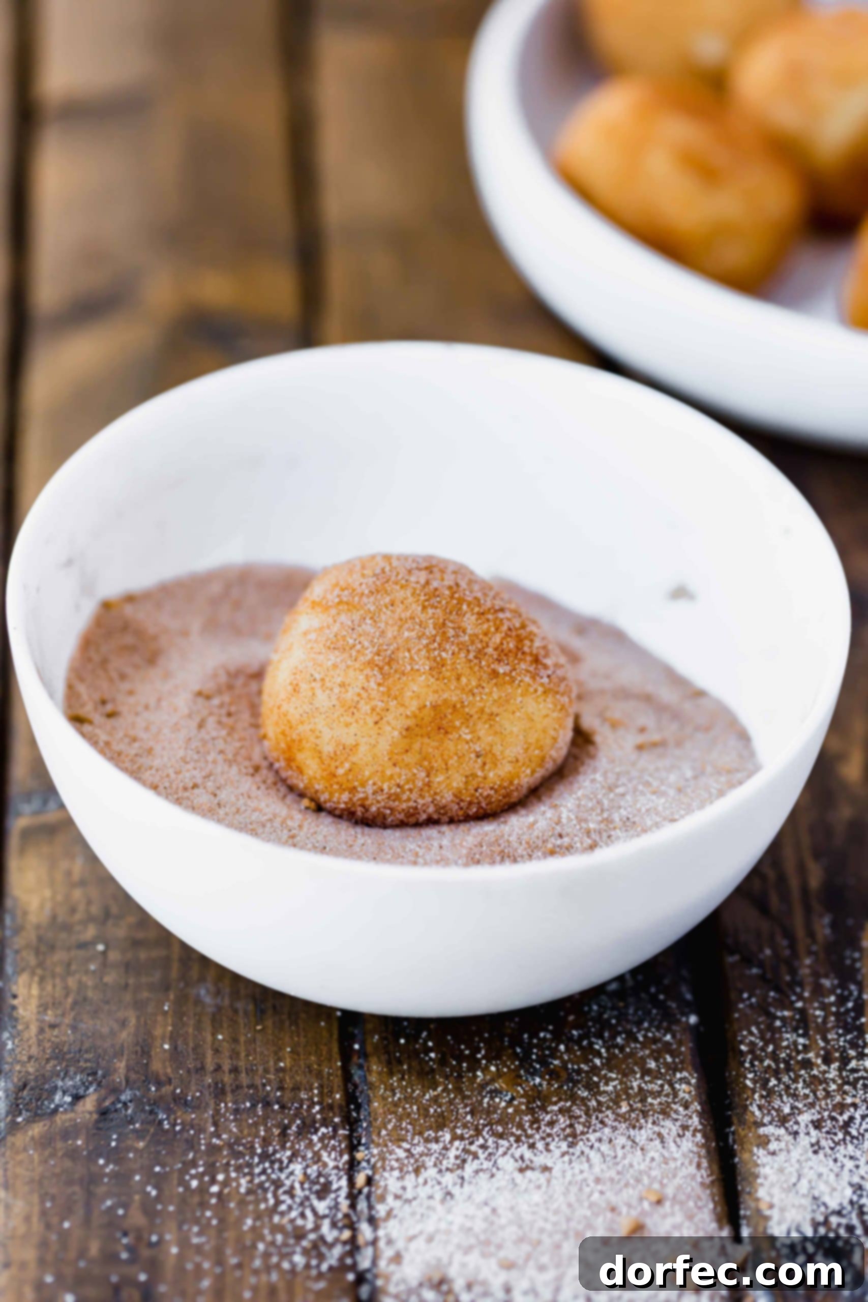 A photo of a ball of snickerdoodle cookie dough sitting in a white bowl, covered in a generous layer of cinnamon sugar, ready for baking.