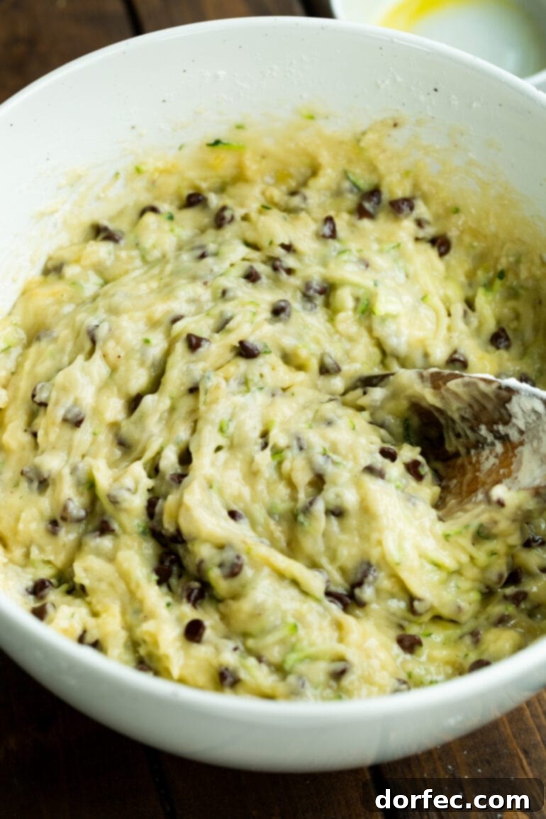 A wooden spoon mixing together the batter for a chocolate chip banana zucchini bread in a white mixing bowl.