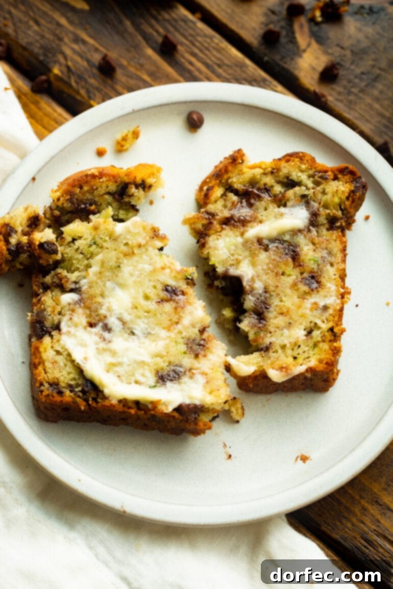 A slice of buttered chocolate chip banana zucchini bread, torn in half and placed on a white dessert plate, ready for a delicious treat.