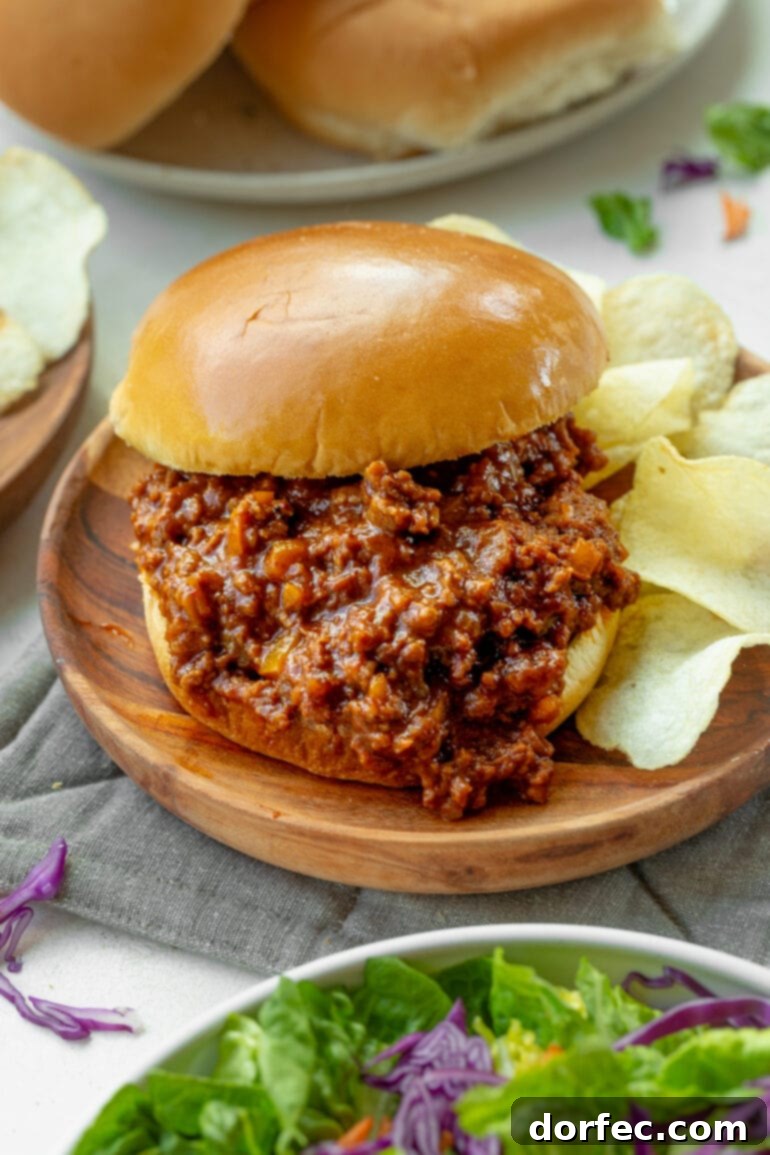 A delicious sloppy joe sandwich, with the savory filling spilling out, resting on a rustic wooden plate next to a stack of crispy potato chips.