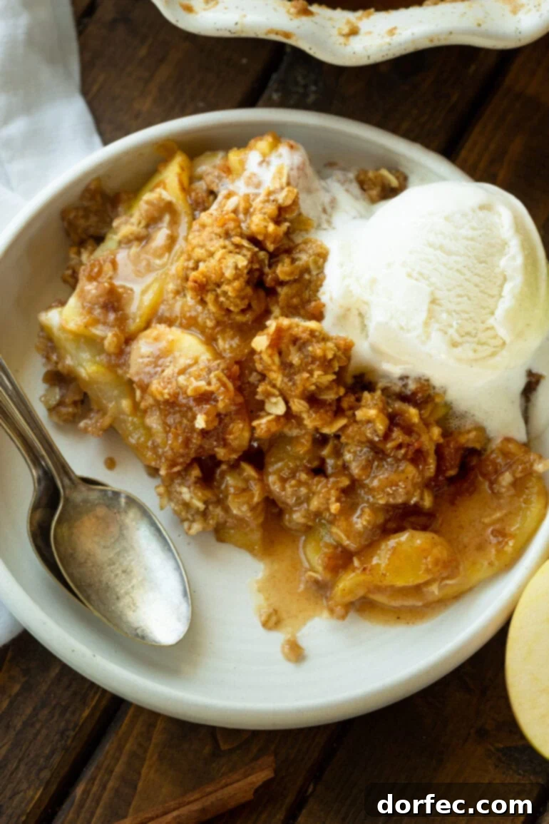A photo of a large serving of warm apple crisp on a white dessert plate topped with a scoop of vanilla ice cream with a spoon sitting on the side, ready to be enjoyed.