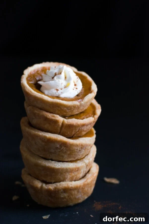 A charming stack of five golden-brown Mini Pumpkin Pies, generously topped with fluffy whipped cream, invitingly presented for a festive occasion.