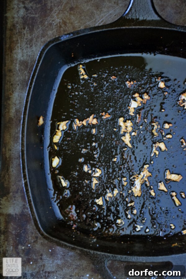 A close-up view of freshly minced garlic sautéing in a cast-iron skillet with a small amount of rendered chicken fat, creating a fragrant base for the herb butter sauce. The garlic pieces are lightly golden, indicating they are perfectly cooked to release their aroma without burning.