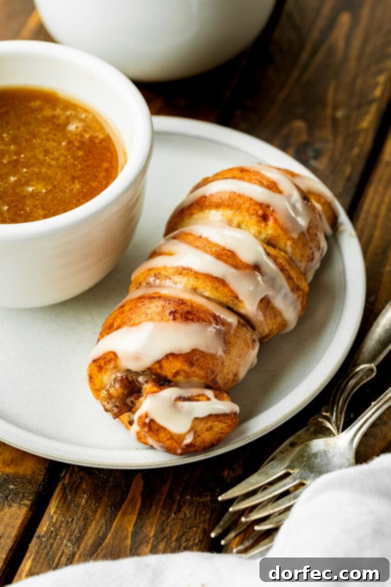 Cinnamon Roll Sausage Poppers 6 A mouth-watering close-up of a single sausage pig in a blanket nestled on a plate, perfectly golden and awaiting a dunk in a small, inviting cup of rich maple butter sauce.