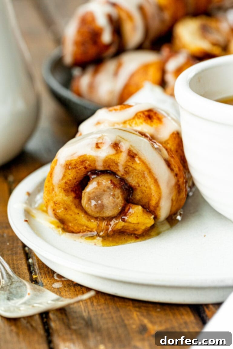Cinnamon Roll Sausage Poppers 7 A single cinnamon roll sausage dunker rests on a plate beside a small bowl of golden maple butter syrup, highlighting the perfect pairing for a sweet and savory breakfast.