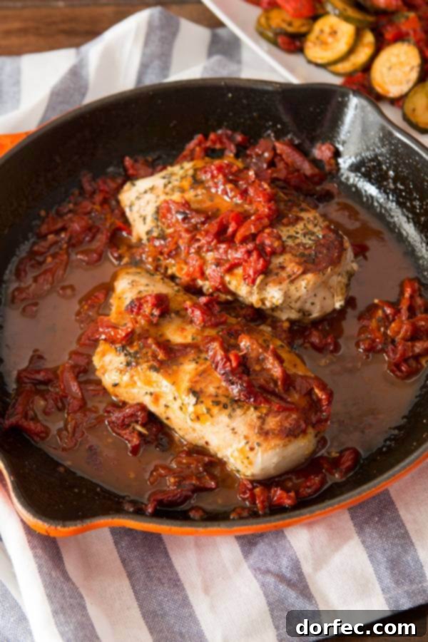 Two cooked chicken breasts, adorned with sun-dried tomatoes, resting in a simple, flavorful sauce within a cast iron skillet.