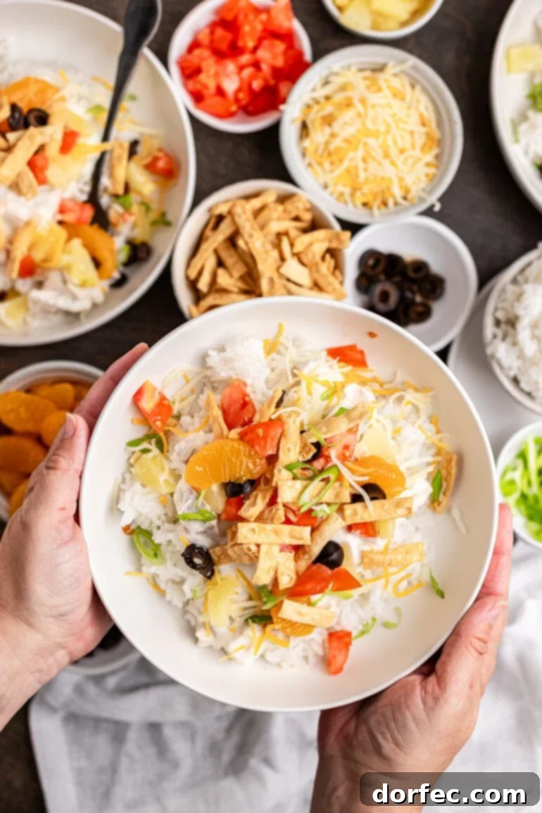 A beautifully arranged white bowl showcasing Hawaiian Haystacks, generously topped with juicy pineapple chunks, grated cheddar cheese, vibrant green onions, and diced fresh tomatoes.