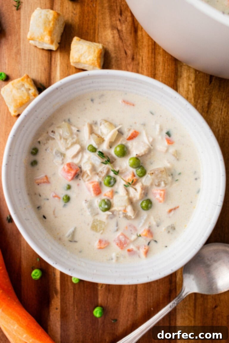 A photo of a bowl of creamy chicken pot pie soup in a white bowl sitting on a wooden table