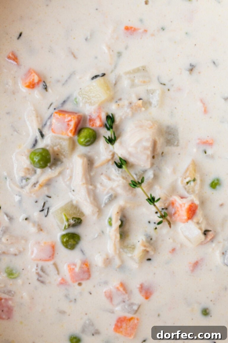 A close up photo taken of creamy chicken pot pie soup with chunks of chicken, peas, carrots and fresh herbs