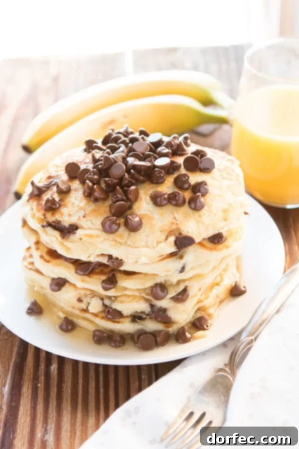 Another enticing stack of chocolate chip pancakes on a white plate, with fresh banana slices and orange juice blurred in the background, hinting at a delightful breakfast spread.