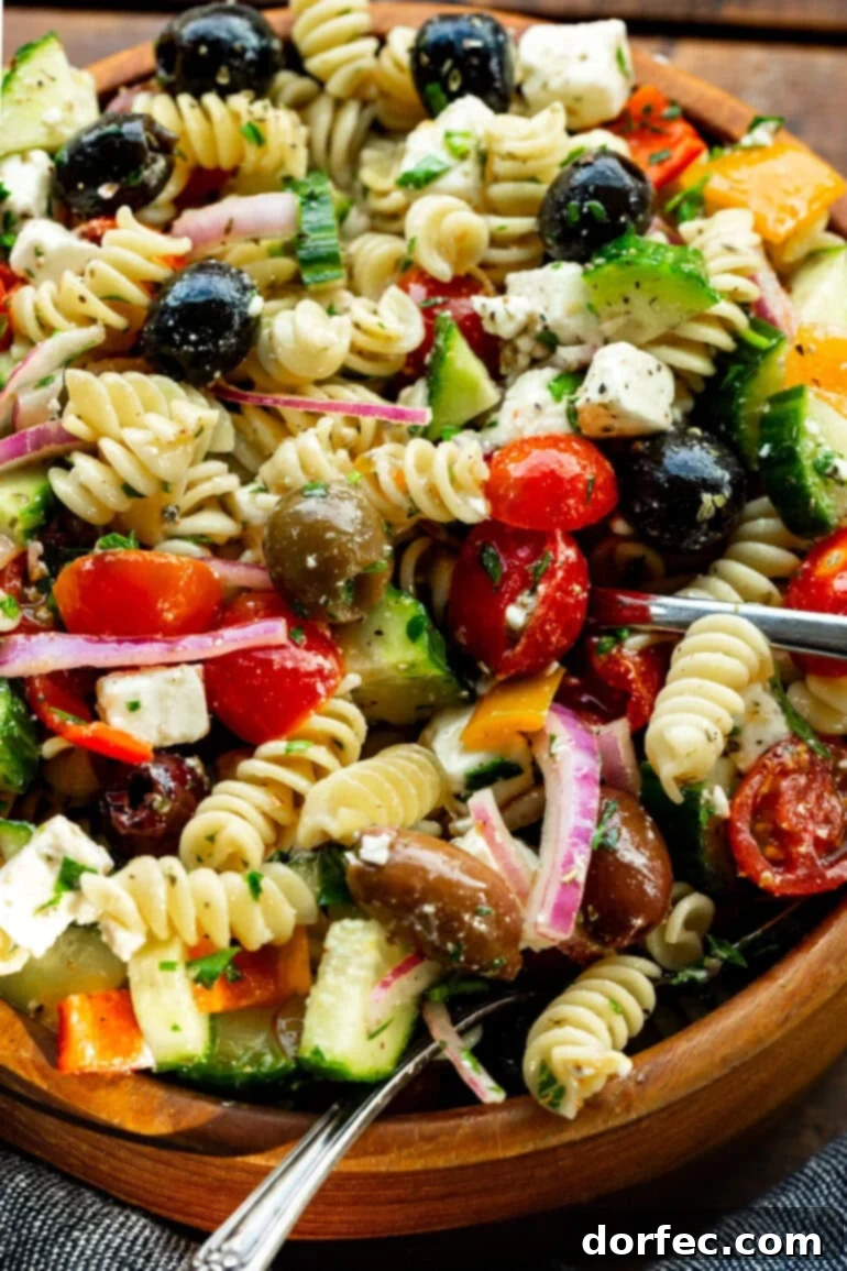 Easy Greek Pasta Salad 2 A large wooden serving bowl overflowing with vibrant Greek pasta salad, featuring curly pasta, red tomatoes, green cucumbers, purple red onions, black olives, and creamy white feta cheese.