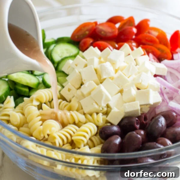 Easy Greek Pasta Salad 11 A hand pouring a stream of vibrant red wine vinaigrette dressing into a bowl filled with Greek pasta salad ingredients.