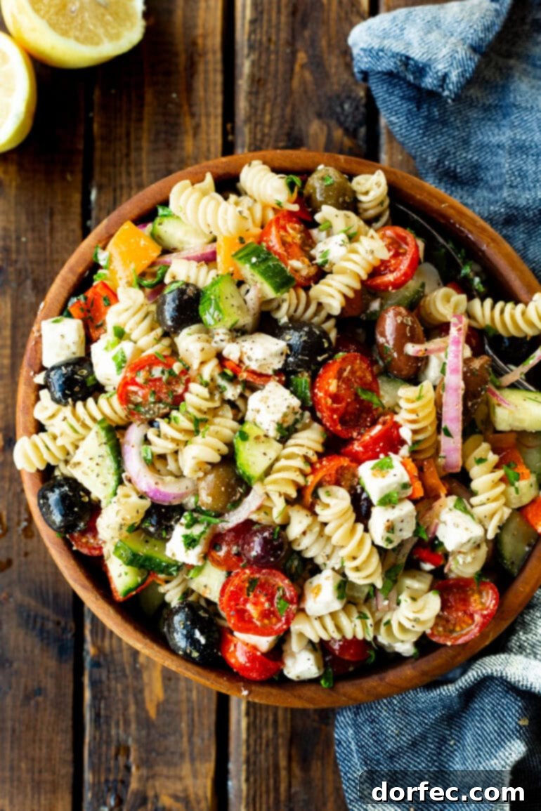 Easy Greek Pasta Salad 5 An overhead shot of a large wooden serving bowl filled with a colorful Greek pasta salad, prominently featuring rotini pasta, bright red tomatoes, crisp green cucumbers, purple red onions, black olives, and creamy feta cheese cubes.