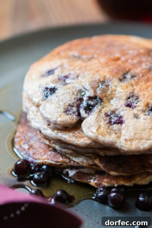 A top-down view of a stack of healthy wild blueberry cottage cheese pancakes on a plate, adorned with fresh berries and a drizzle of syrup, emphasizing a gluten-free breakfast option.