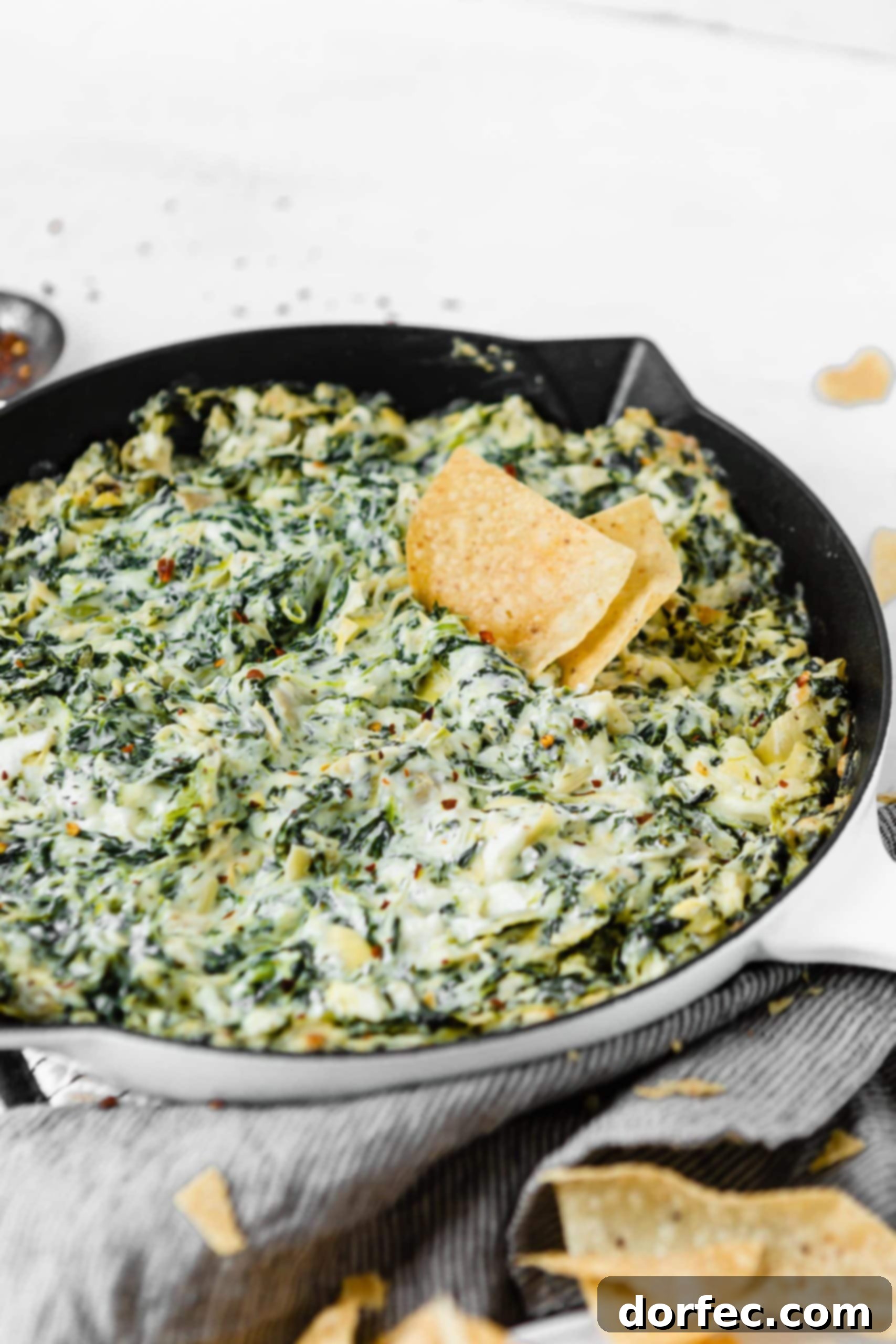 Cheesy Spinach Artichoke Dip A cast iron skillet filled with spinach and artichoke dip. You can see the creamy white cheese sauce with green spinach and artichokes. There are two tortilla chips in the dip.