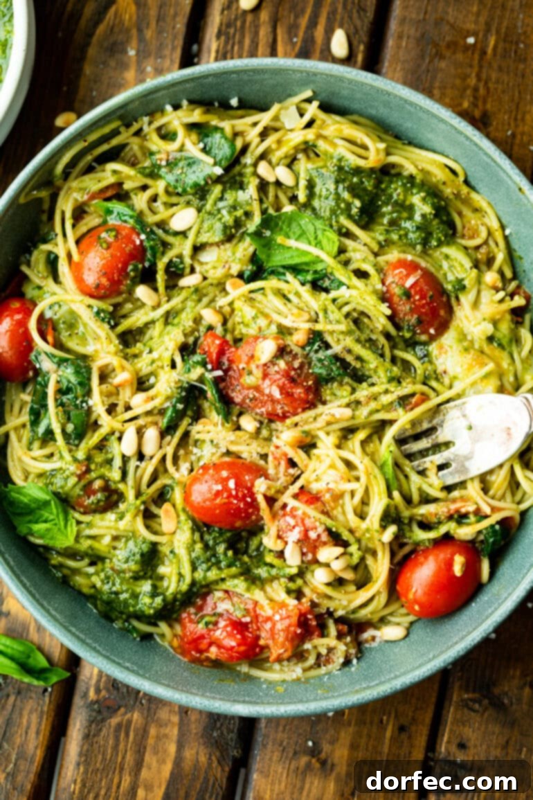 A vibrant photo of cooked angel hair pasta generously tossed with fresh basil pesto, bright red grape tomatoes, crunchy pine nuts, delicate fresh basil leaves, and grated Parmesan cheese, ready to be served.