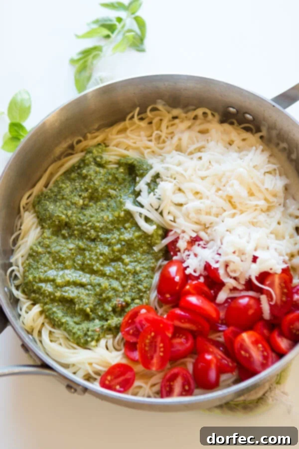Fresh basil pesto, shredded mozzarella cheese, delicate angel hair pasta, and vibrant grape tomatoes arranged in a large skillet, representing the core ingredients before mixing into a delicious pasta dish.