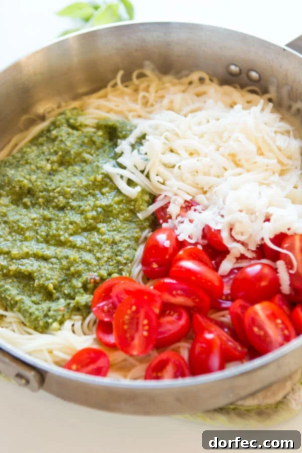 A close-up view of fresh basil pesto, shredded mozzarella cheese, cooked pasta, and colorful grape tomatoes nestled together in a skillet, capturing the rich textures and preparation stage of the meal.