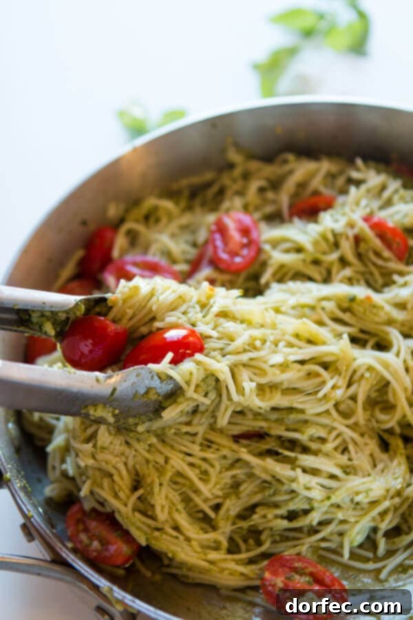 Tongs lifting a savory serving of basil pesto pasta from a skillet, showing the perfectly coated noodles and melted cheese, ready for plating.