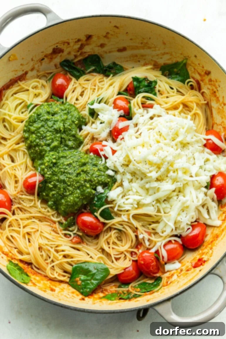 A large pot of cooked angel hair pasta forms the base, topped with a generous pile of vibrant green pesto sauce, shredded mozzarella cheese, fresh basil leaves, and whole grape tomatoes, showcasing the main components before mixing.