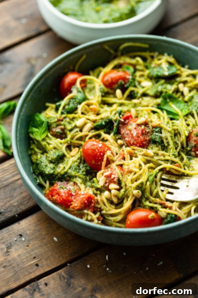 A large bowl brimming with cooked angel hair pasta, artfully tossed with fresh basil pesto, plump grape tomatoes, fragrant basil leaves, shredded mozzarella cheese, and crunchy pine nuts, creating a mouth-watering ensemble.