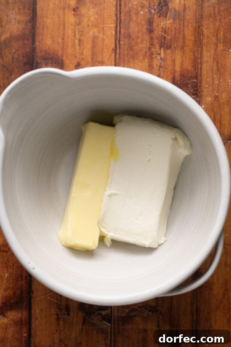 Close-up of softened cream cheese and butter being beaten together in a large bowl with a hand mixer, starting the process of making smooth chocolate cream cheese frosting.